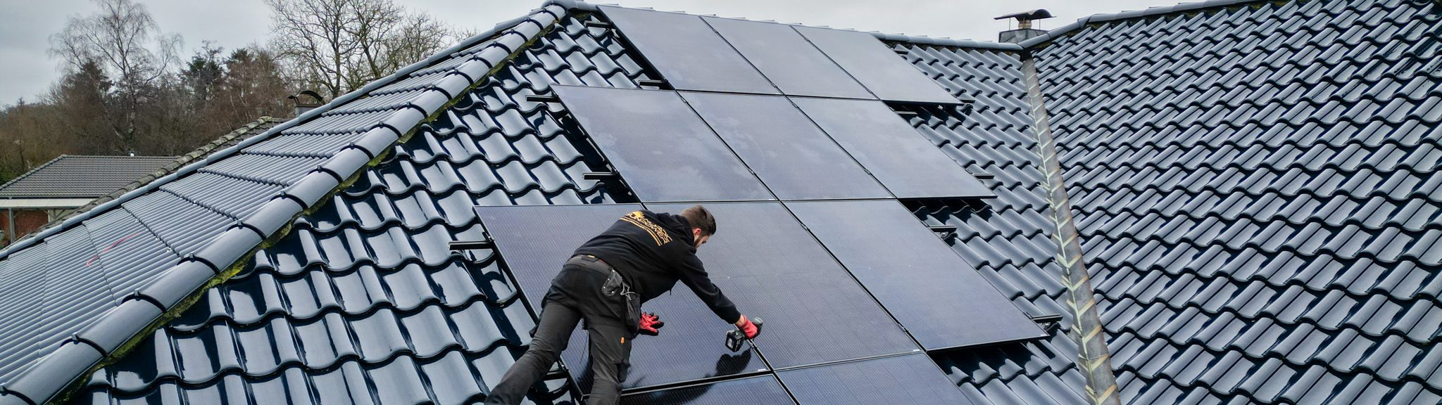 Zwei Monteure installieren schwarze Solarmodule auf einem dunkel glasierten Ziegeldach. Einer der Handwerker arbeitet mit einem Akkuschrauber, der andere steht auf dem Gerüst; der Himmel ist bewölkt.