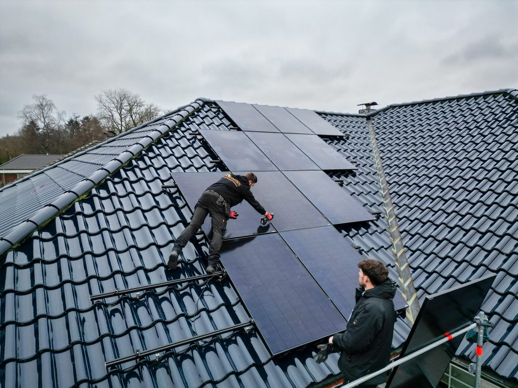 Zwei Monteure installieren schwarze Solarmodule auf einem dunkel glasierten Ziegeldach. Einer der Handwerker arbeitet mit einem Akkuschrauber, der andere steht auf dem Gerüst; der Himmel ist bewölkt.
