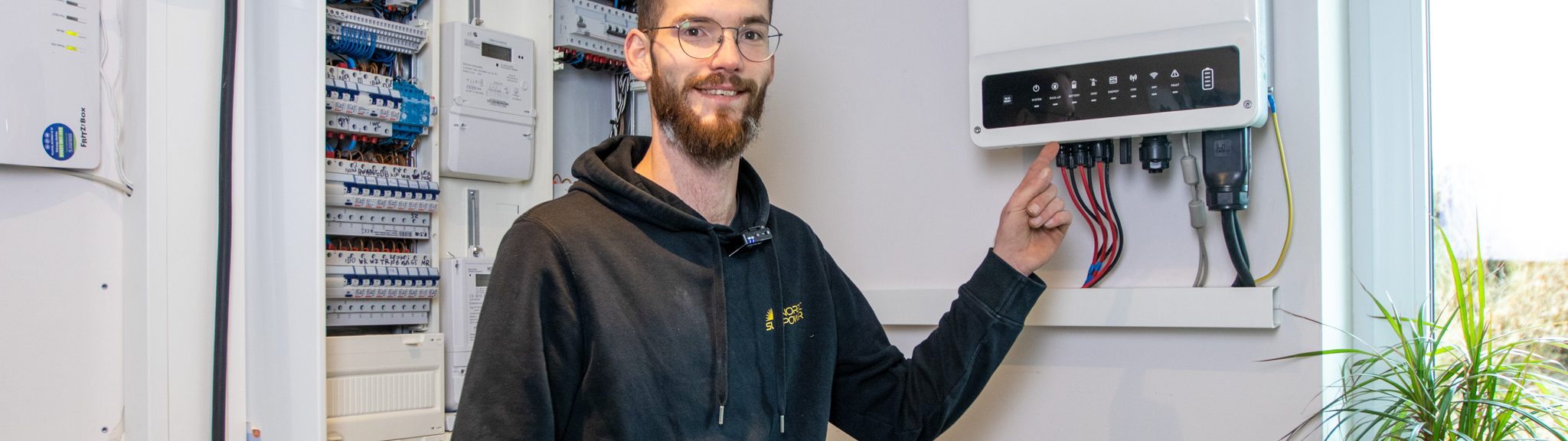 Elektriker steht neben einem montierten Wechselrichter an der Wand und zeigt auf das Display. Im Hintergrund sind geöffnete Sicherungskästen und Kabel zu sehen, rechts ein Fenster mit einer Zimmerpflanze.