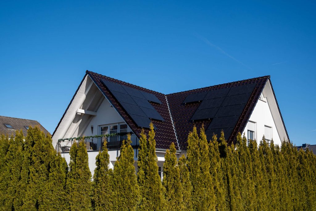 Modernes Einfamilienhaus mit dunkelbraunem Satteldach und Photovoltaikanlage auf beiden Dachseiten. Im Vordergrund ist eine dichte Hecke zu sehen, darüber blauer Himmel ohne Wolken.