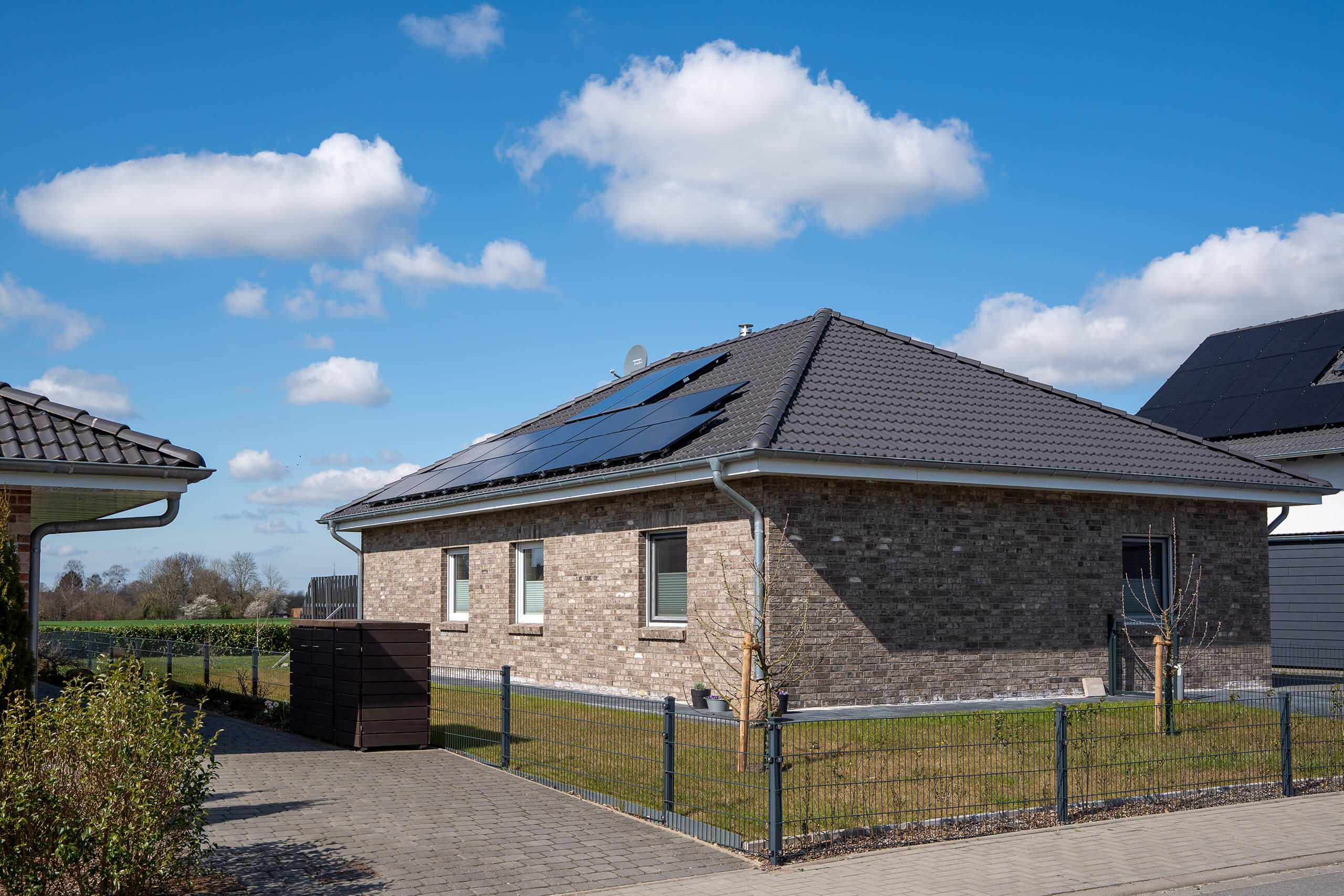 Einfamilienhaus mit grauem Dach und installierter Photovoltaikanlage auf der Südseite. Im Vordergrund sind Garten, Zaun und gepflasterte Einfahrt zu sehen, bei sonnigem Wetter mit blauem Himmel und weißen Wolken.
