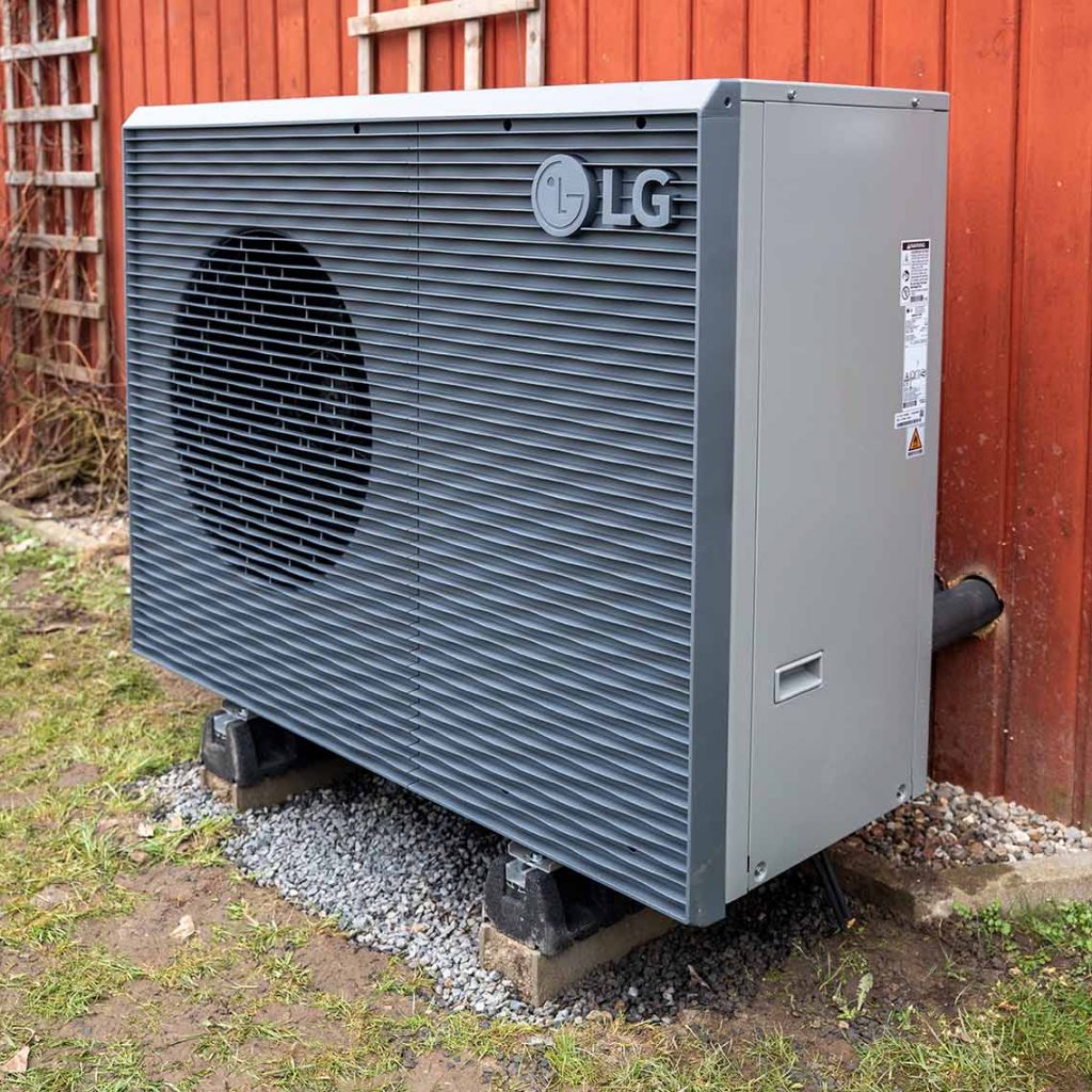 Außeneinheit einer Wärmepumpe der Marke LG steht auf Fundamentsteinen vor einer roten Holzfassade. Das Gerät ist grau und über Leitungen mit dem Gebäude verbunden.