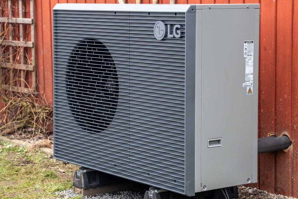 Außeneinheit einer Wärmepumpe der Marke LG steht auf Fundamentsteinen vor einer roten Holzfassade. Das Gerät ist grau und über Leitungen mit dem Gebäude verbunden.