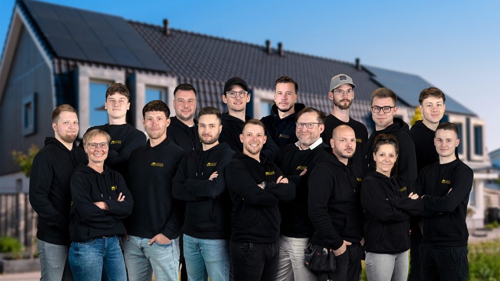 Teamfoto von Nordic Sunpower vor einem Haus mit Solaranlage. Die Mitarbeiterinnen und Mitarbeiter tragen schwarze Firmenkleidung mit Logo und stehen gemeinsam vor einem sonnigen Hintergrund.