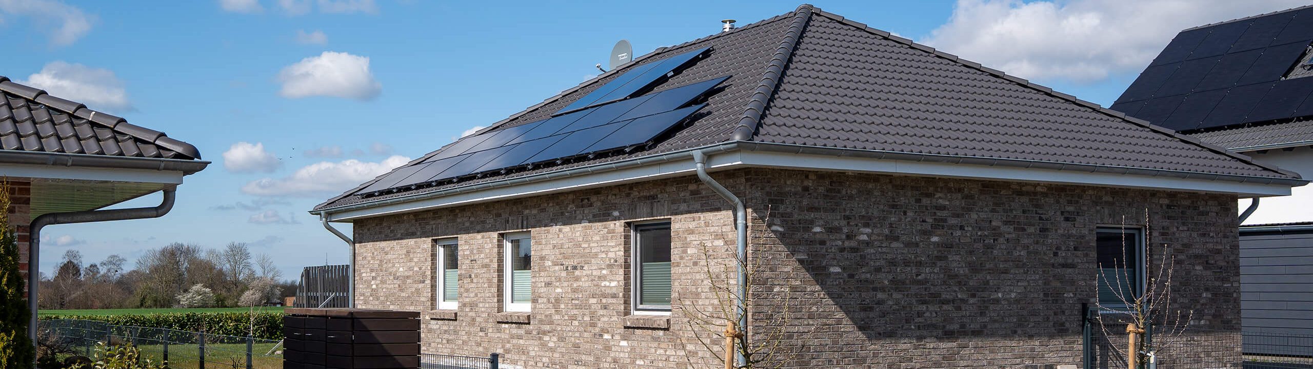 Einfamilienhaus mit Klinkerfassade und dunkelgrauem Walmdach, auf dem eine Photovoltaikanlage installiert ist. Im Hintergrund sind Nachbarhäuser, Wiesen und blauer Himmel mit einzelnen Wolken zu sehen.