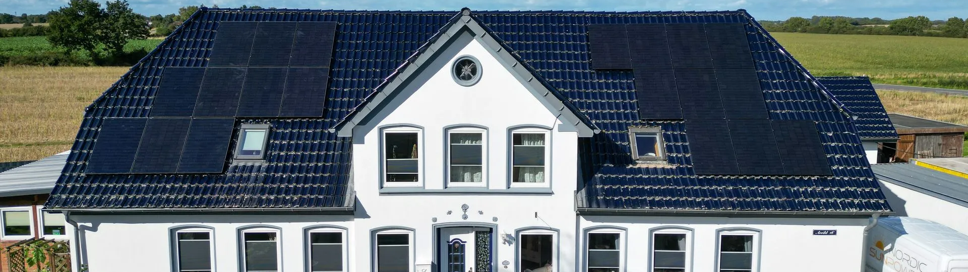 Großes weißes Wohnhaus mit dunkelblauem Dach und umfangreicher Photovoltaikanlage auf beiden Dachseiten. Im Hintergrund sind Felder und blauer Himmel zu sehen.