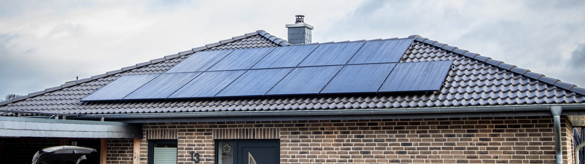 Einfamilienhaus mit hellbrauner Klinkerfassade und grauem Walmdach, auf dem eine Photovoltaikanlage installiert ist. Der Himmel ist leicht bewölkt.
