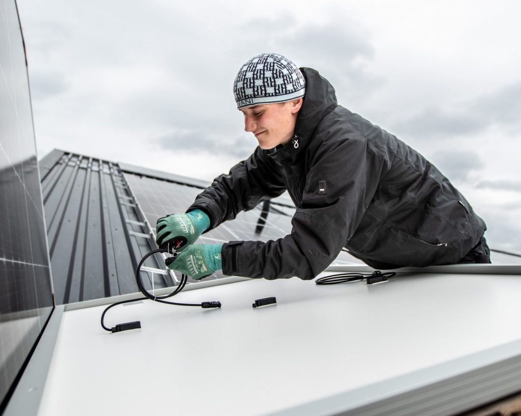 Handwerker montiert Solarmodule auf einem Metalldach und schließt Kabelverbindungen an. Er trägt wetterfeste Arbeitskleidung, Handschuhe und eine Mütze; der Himmel ist bewölkt.