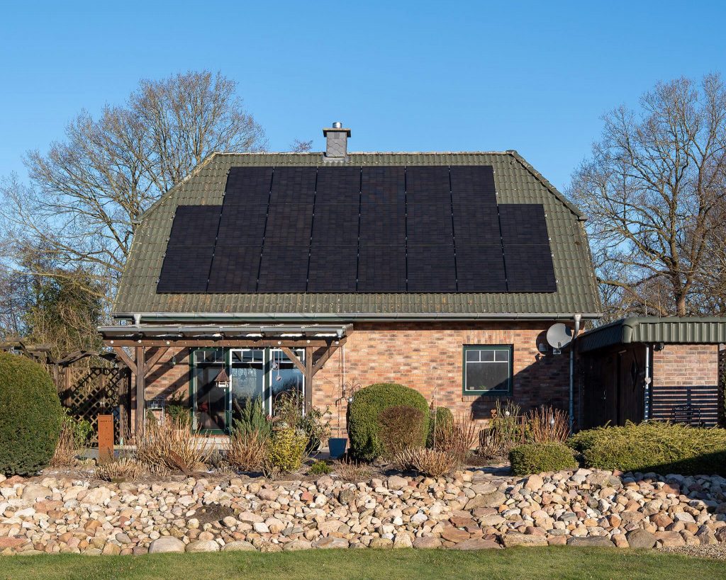 Einfamilienhaus mit roter Klinkerfassade und grünlichem Satteldach, auf dem eine großflächige Photovoltaikanlage installiert ist. Vor dem Haus befinden sich Gartenbeete, Sträucher und eine niedrige Steinmauer.