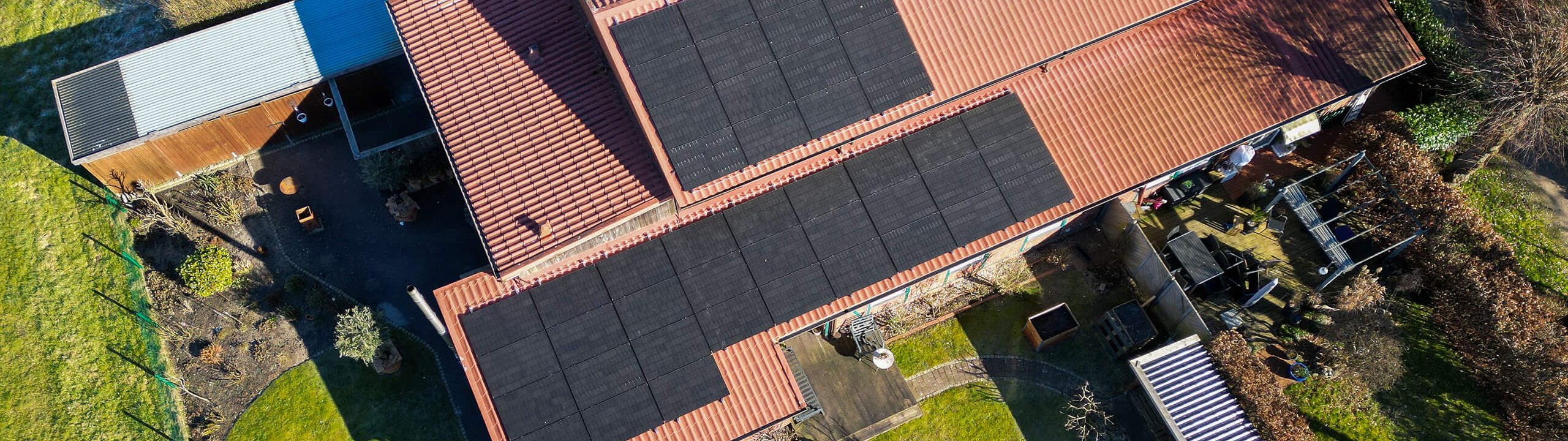 Luftaufnahme eines langgestreckten Wohnhauses mit rotem Ziegeldach und mehreren Reihen schwarzer Photovoltaikmodule. Rund um das Gebäude liegen Gartenflächen, Nebengebäude und ein kleiner Hofbereich.