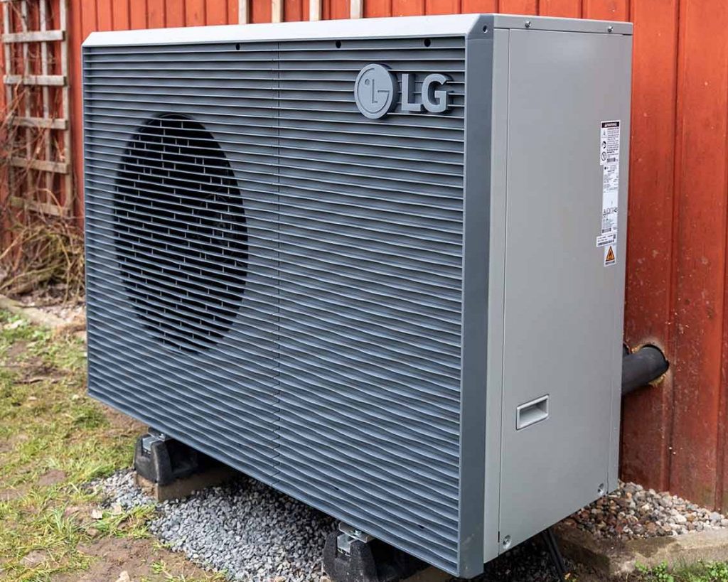 Außeneinheit einer Wärmepumpe der Marke LG steht auf Fundamentsteinen vor einer roten Holzfassade. Das Gerät ist grau und über Leitungen mit dem Gebäude verbunden.