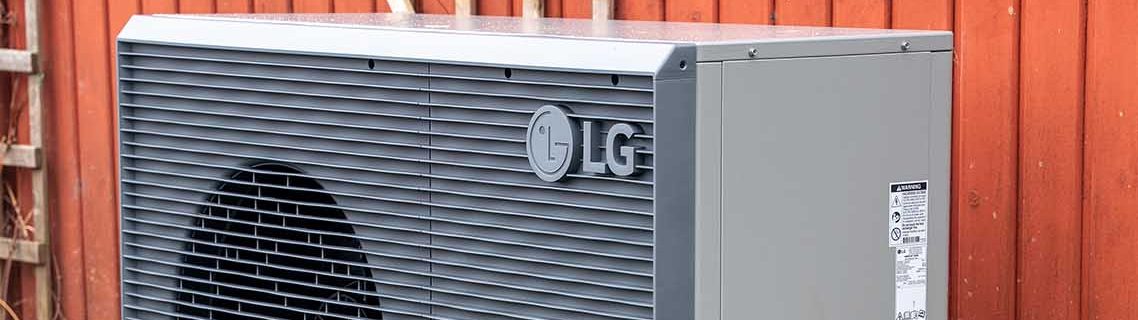 Außeneinheit einer Wärmepumpe der Marke LG steht auf Fundamentsteinen vor einer roten Holzfassade. Das Gerät ist grau und über Leitungen mit dem Gebäude verbunden.