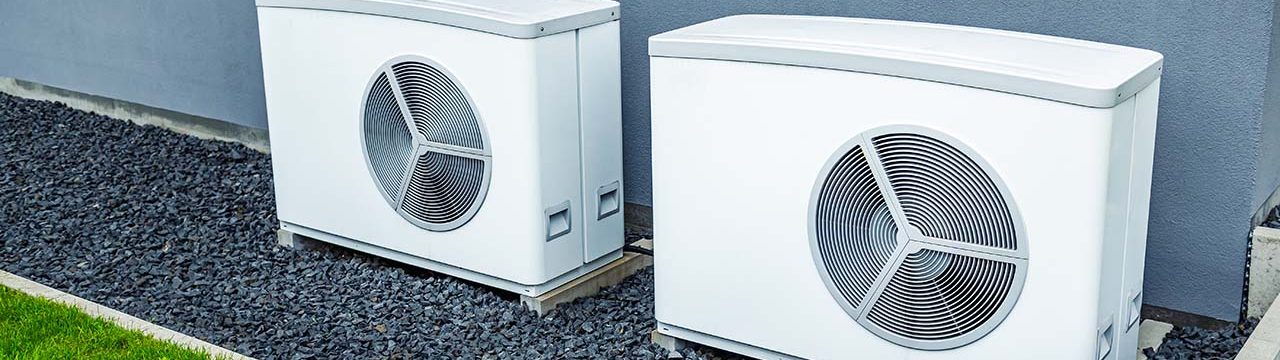 Zwei weiße Außeneinheiten von Wärmepumpen stehen nebeneinander vor einer grauen Hauswand auf einem mit dunklem Kies bedeckten Untergrund. Links im Bild ist eine Rasenfläche zu sehen.