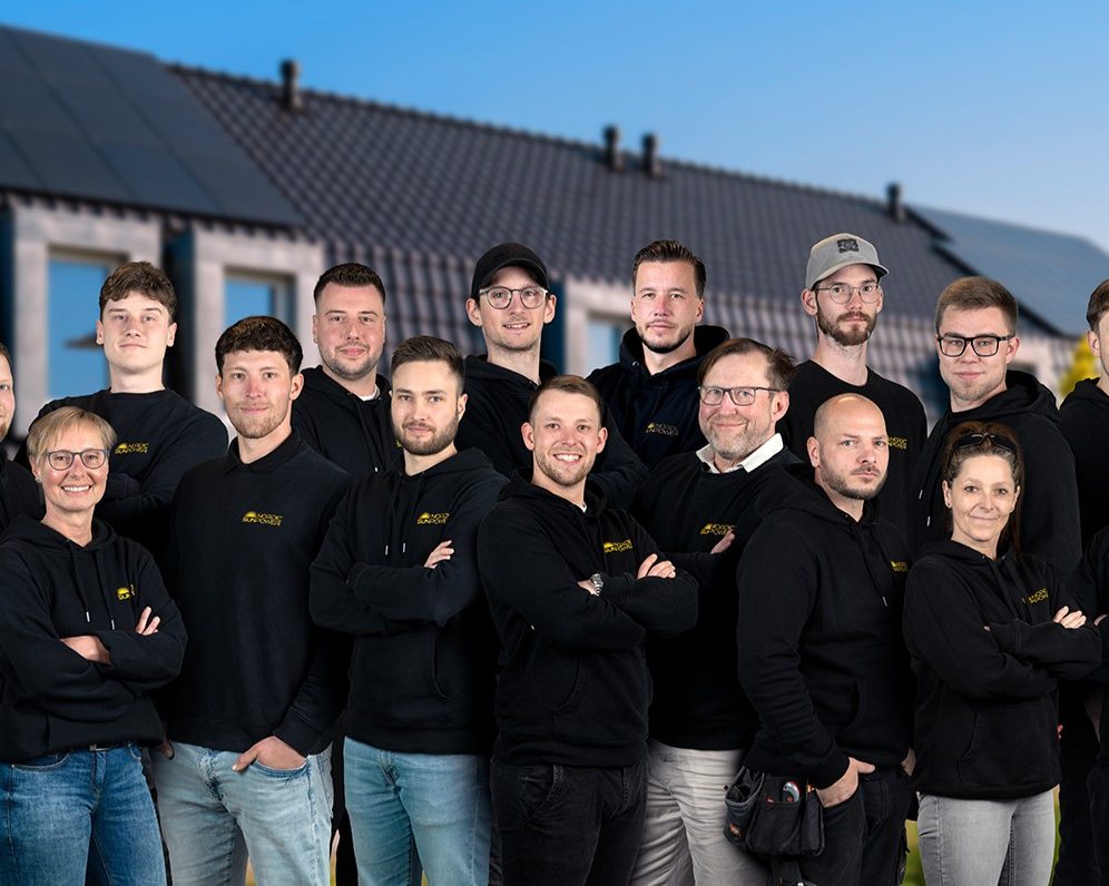 Teamfoto von Nordic Sunpower vor einem Haus mit Solaranlage. Die Mitarbeiterinnen und Mitarbeiter tragen schwarze Firmenkleidung mit Logo und stehen gemeinsam vor einem sonnigen Hintergrund.